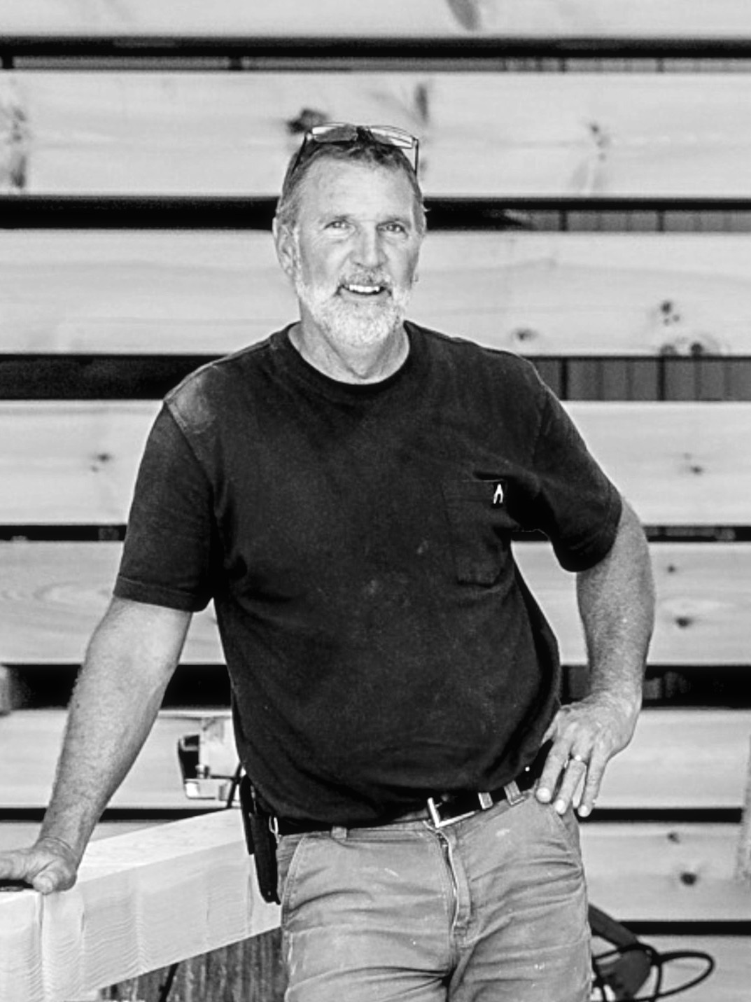 Greg Sickler in the shop, leaning on a timber in front of a stack of beams, black and white