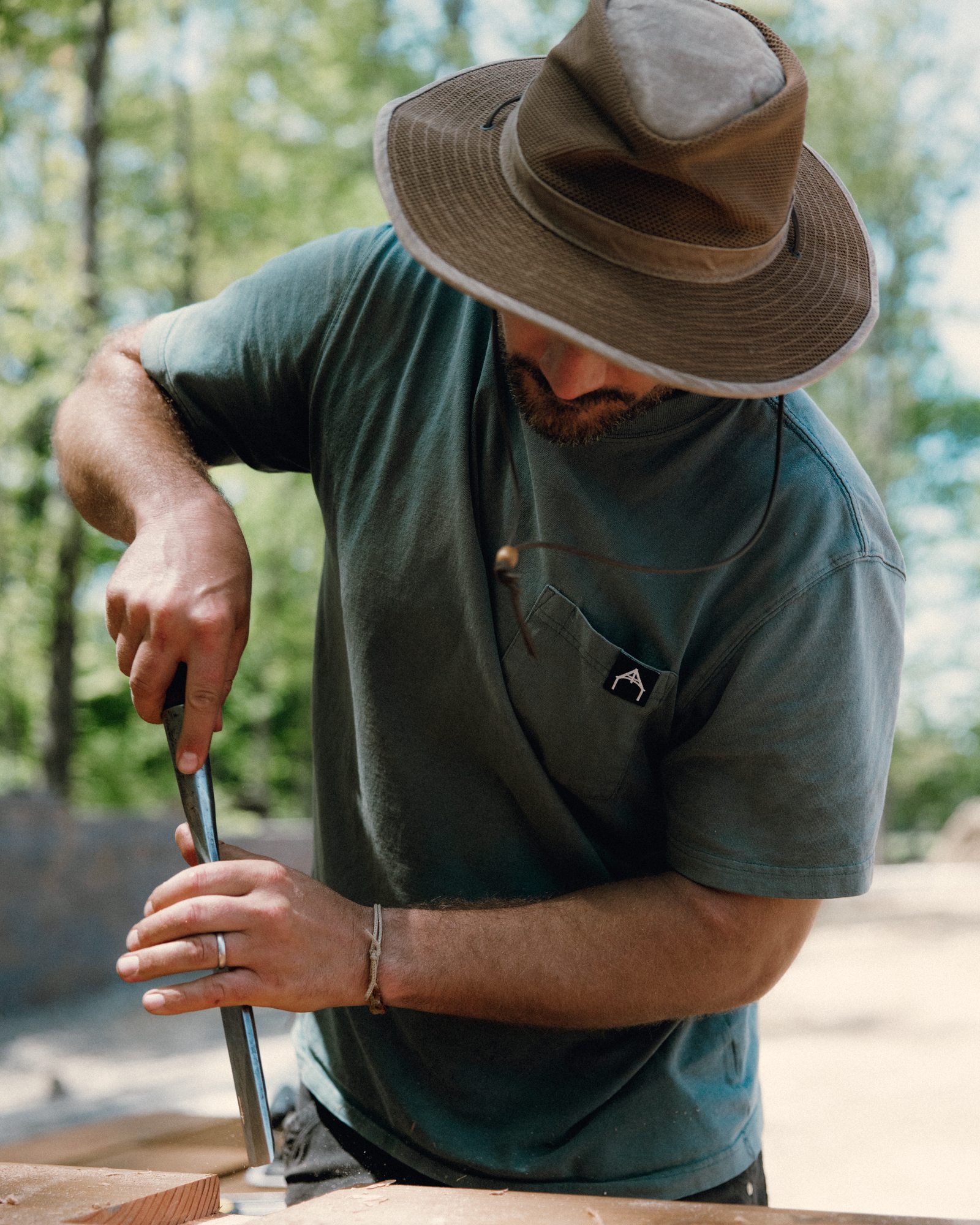 Timber framer paring a mortise with a framing chisel on site, trees behind him