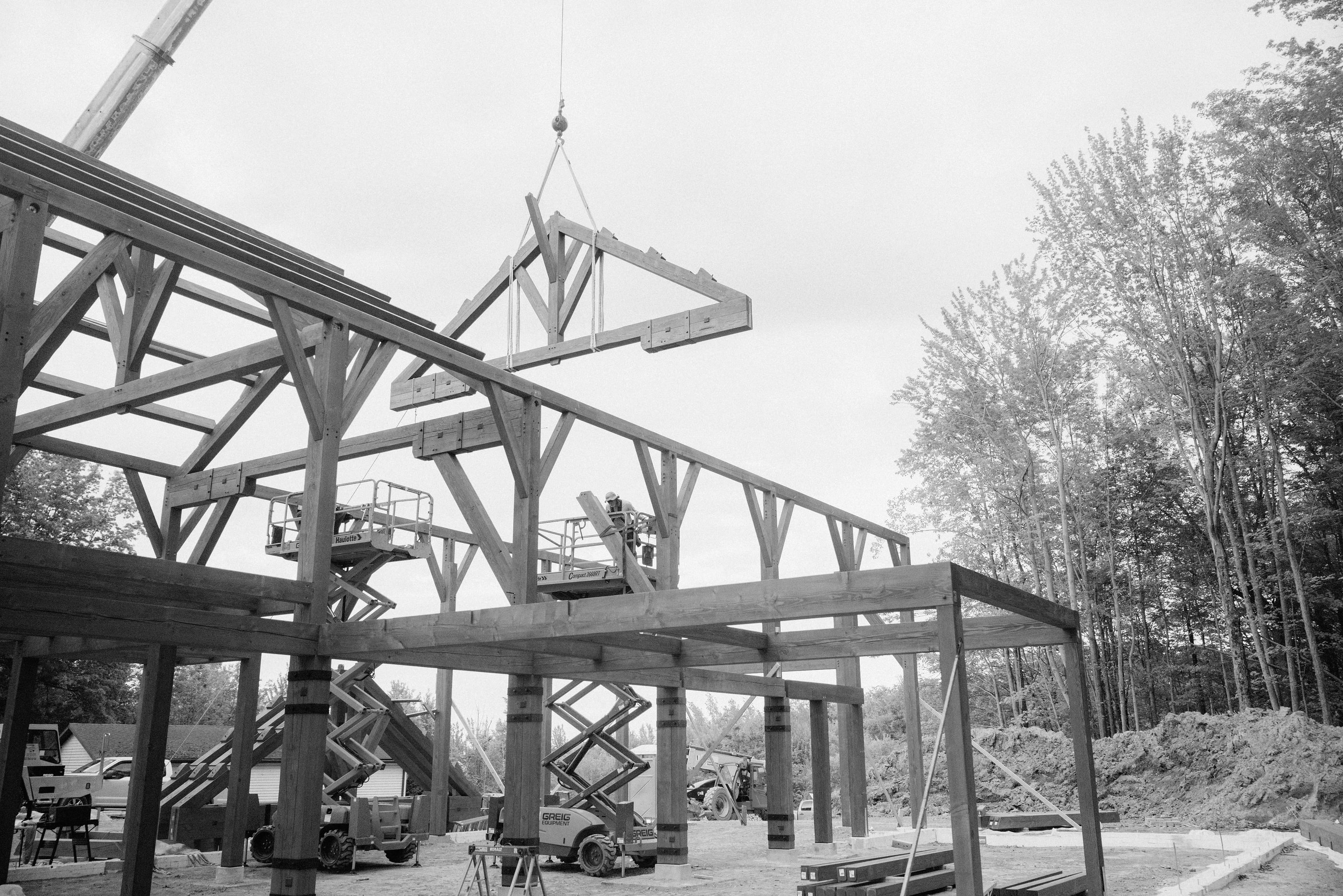 A crane lifting a truss section into the raised frame, crew working from scissor lifts below, black and white