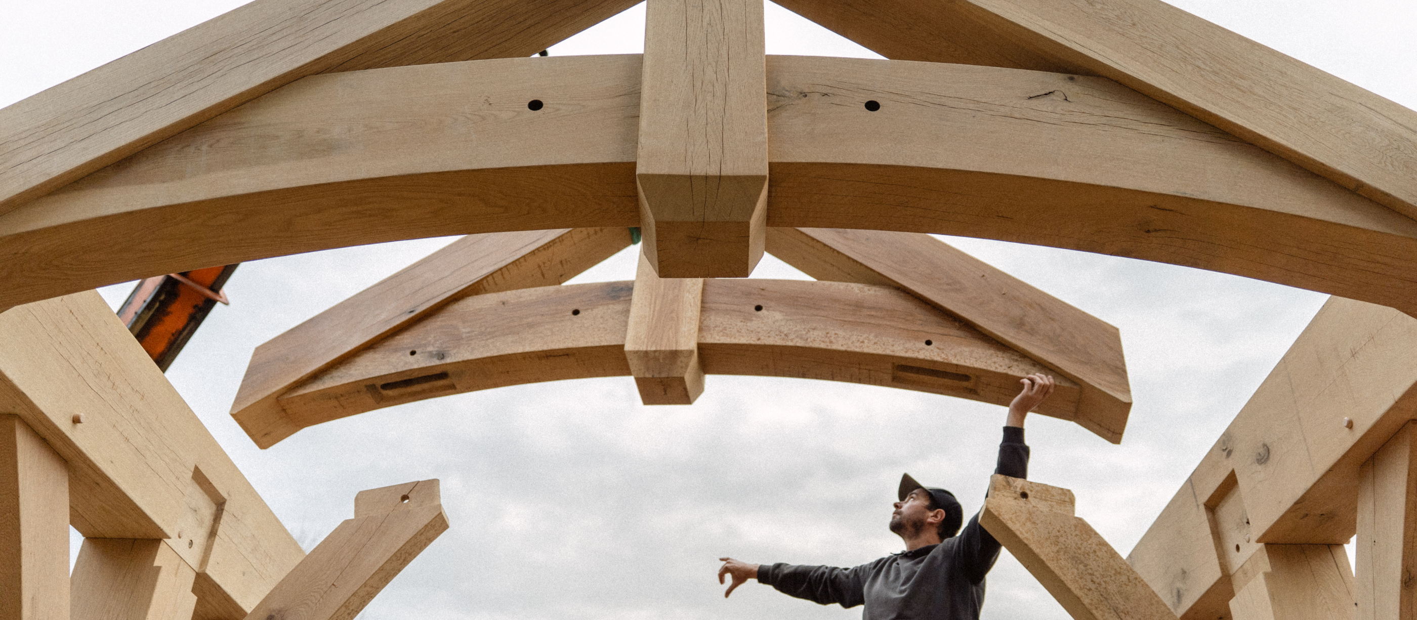 Crew member on a lift guiding a curved oak tie into a truss during a raising, crane boom behind