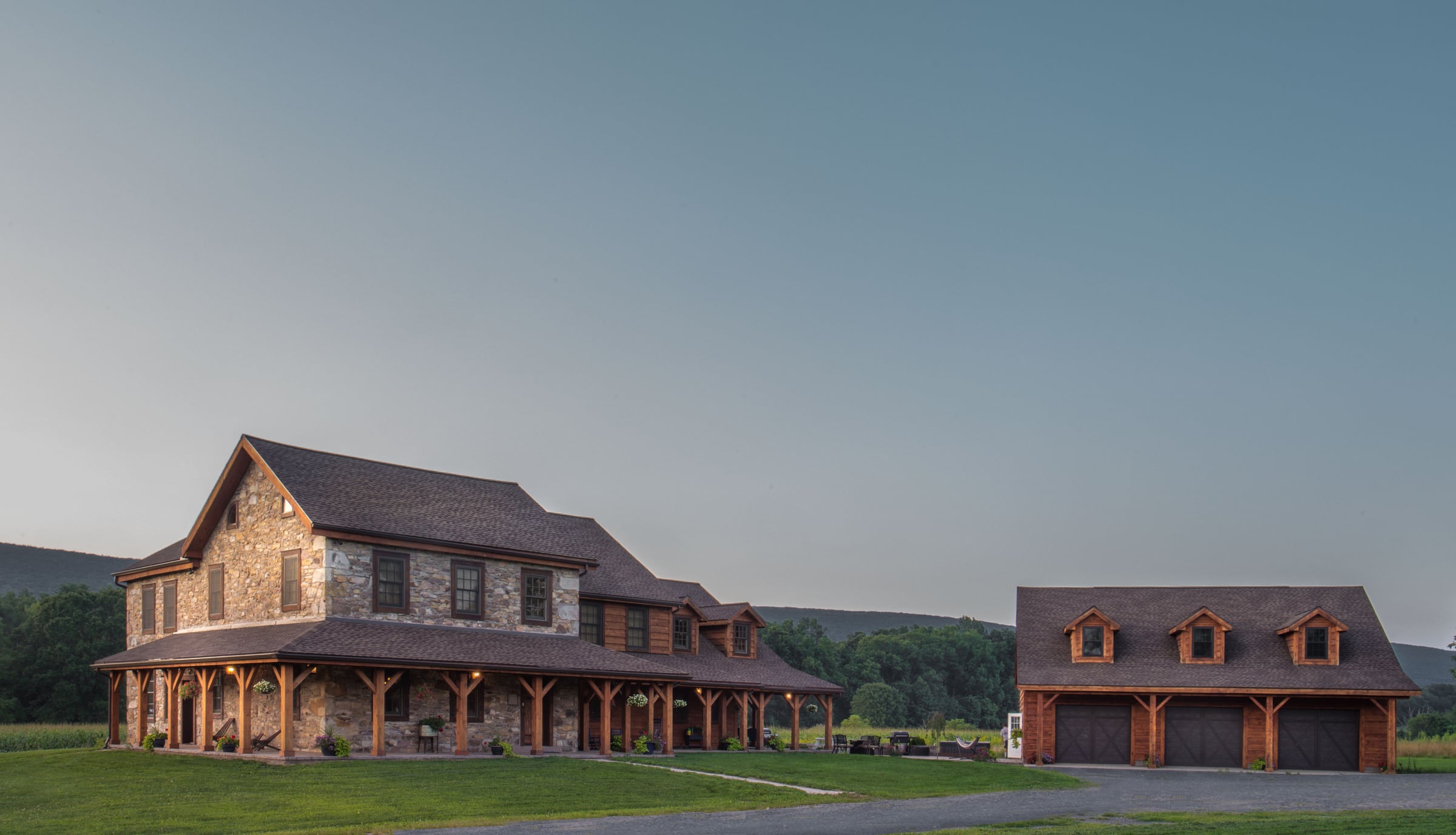 The stone house, the timber frame addition and the garage at dusk, the wraparound porch lit, hills behind