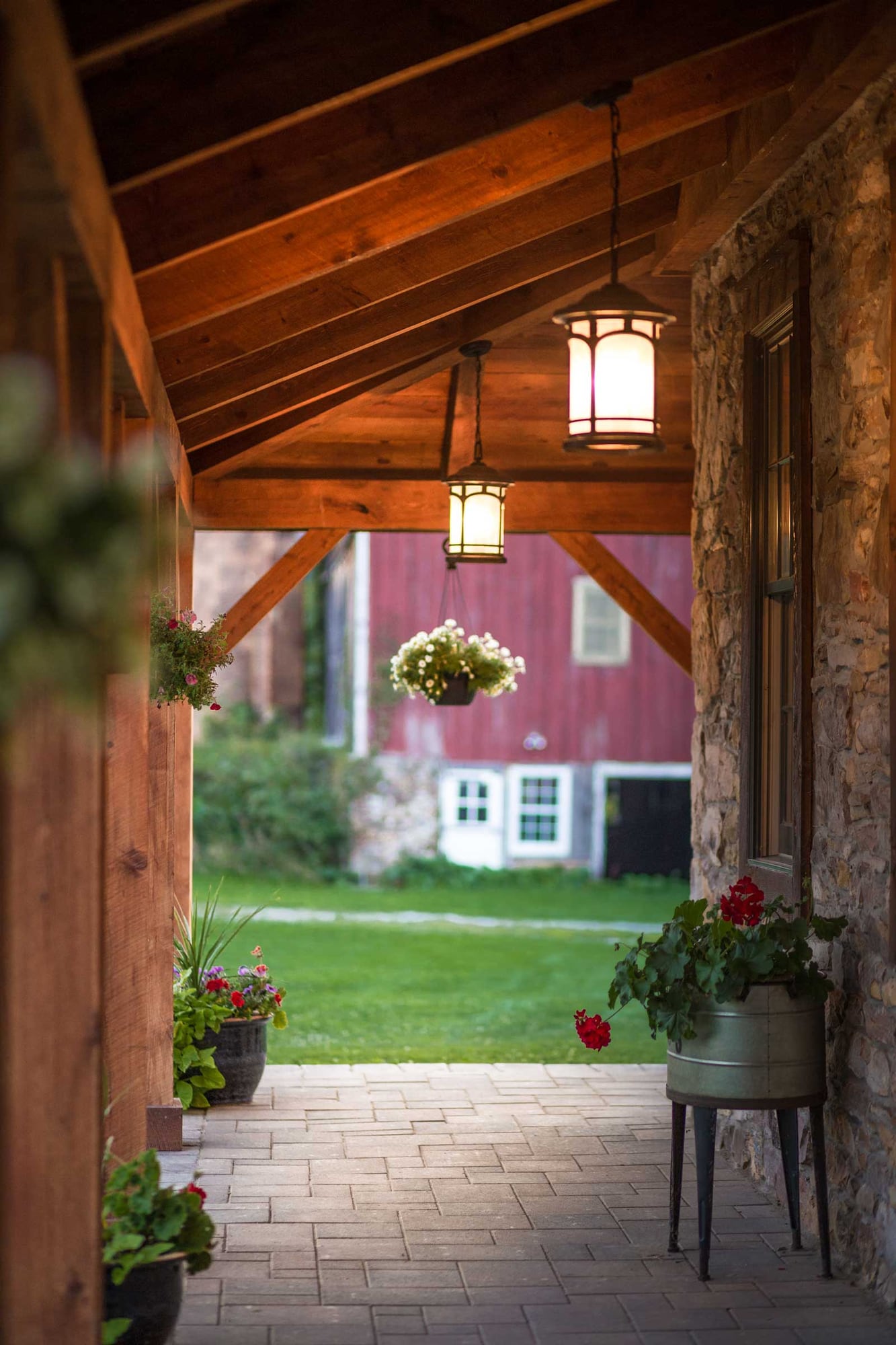 Down the porch toward the red barn, lanterns and hanging flowers under the hemlock rafters
