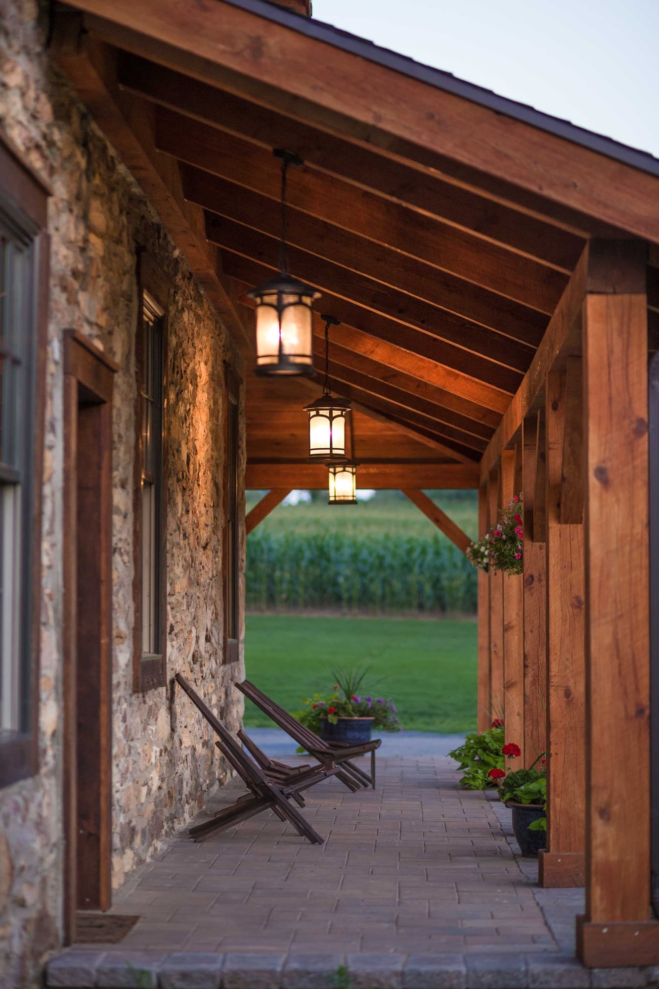 The porch at dusk: hemlock posts and rafters, lanterns lit, chairs against the stone wall, a cornfield beyond