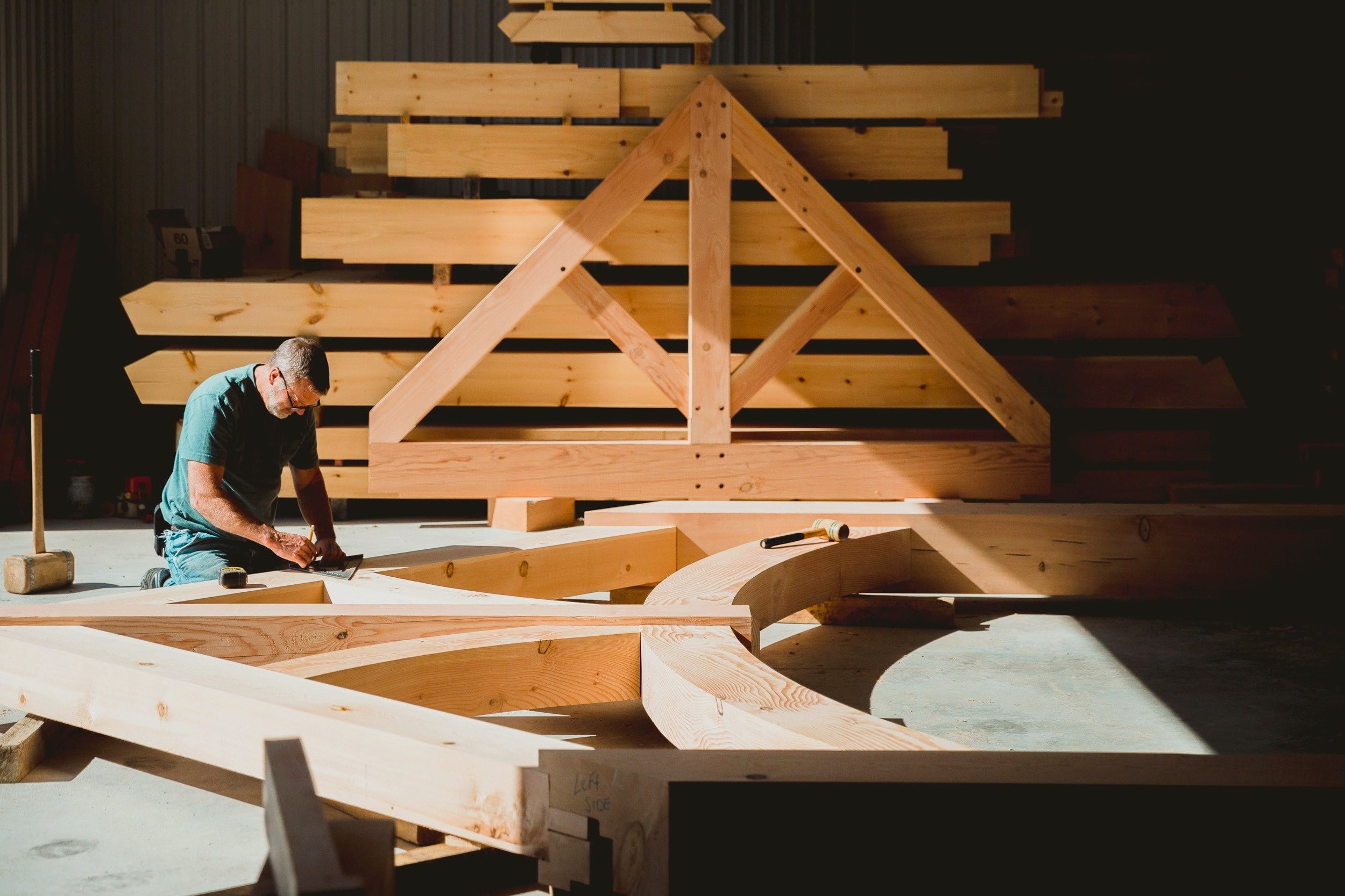 A framer kneels to mark out a curved truss on the shop floor, a finished truss and stacked timbers behind him
