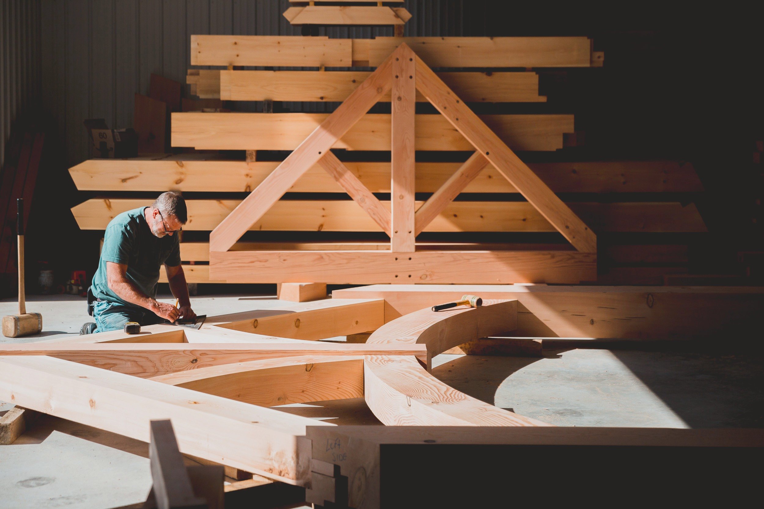 A framer kneels to mark out a curved truss on the shop floor, a finished truss and stacked timbers behind him