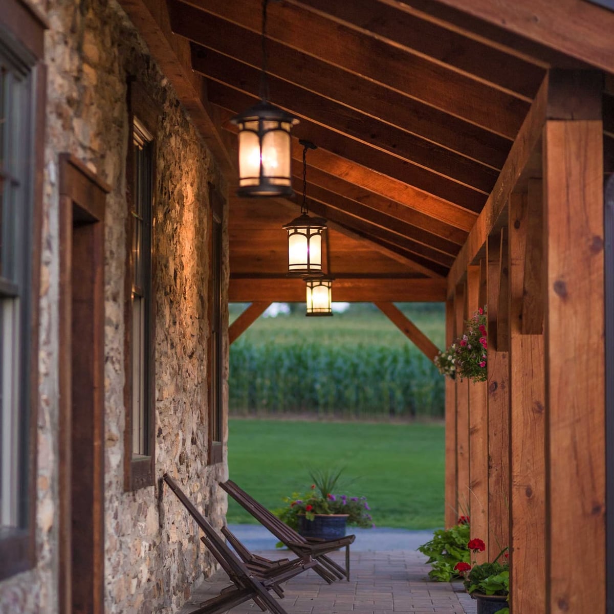 The stone house porch at dusk: hemlock posts and rafters, lanterns lit, chairs against the stone wall, a cornfield beyond