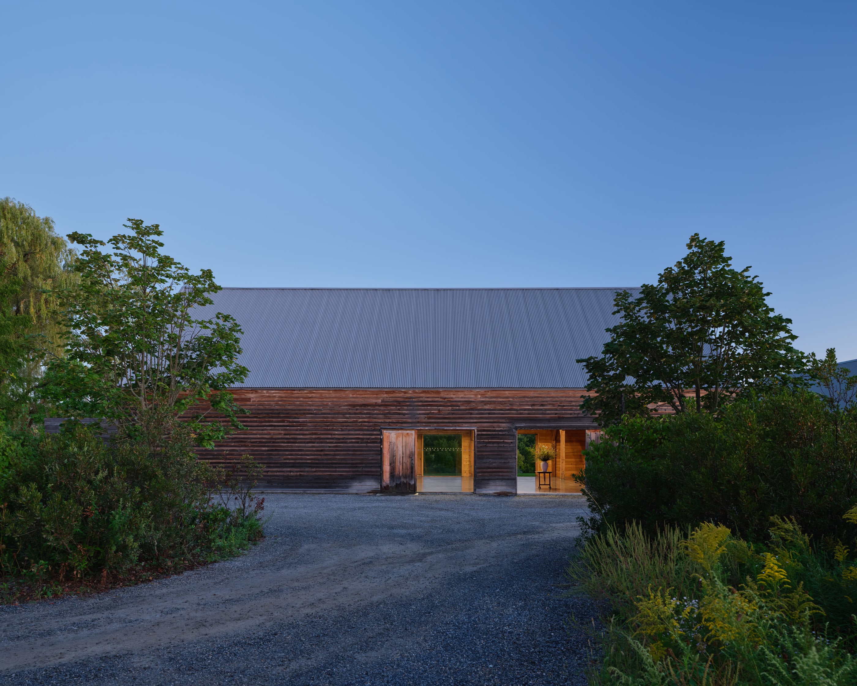 The INNESS barn at dusk, its doors open and the interior lit, gravel drive and trees in the blue hour