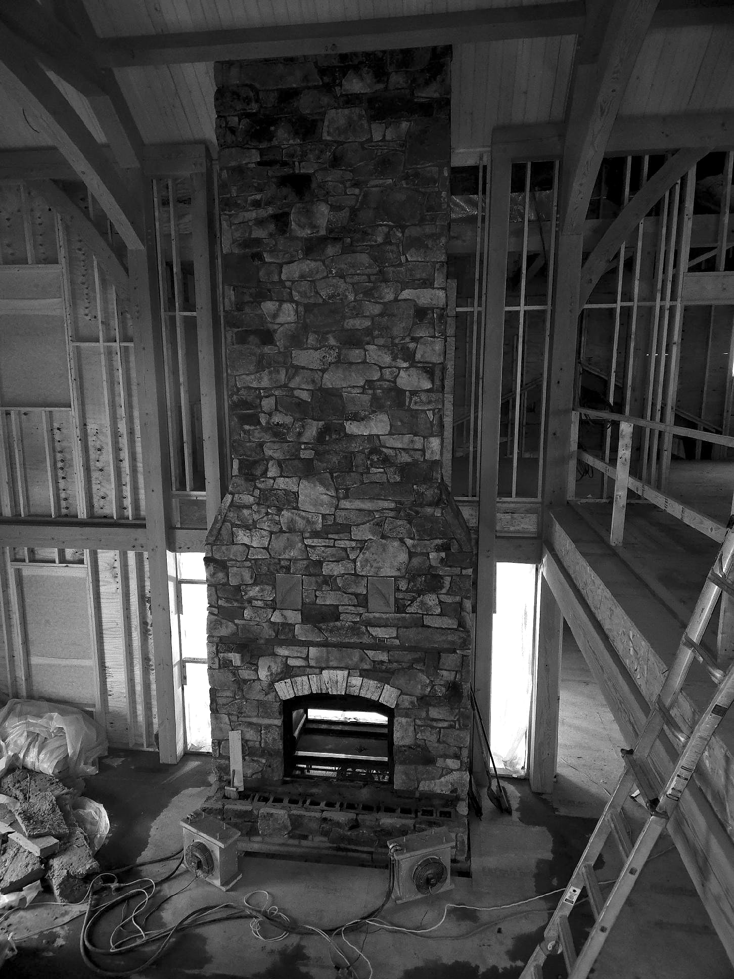 The stone chimney rising through the great room between the frame’s arched braces, under construction, black and white