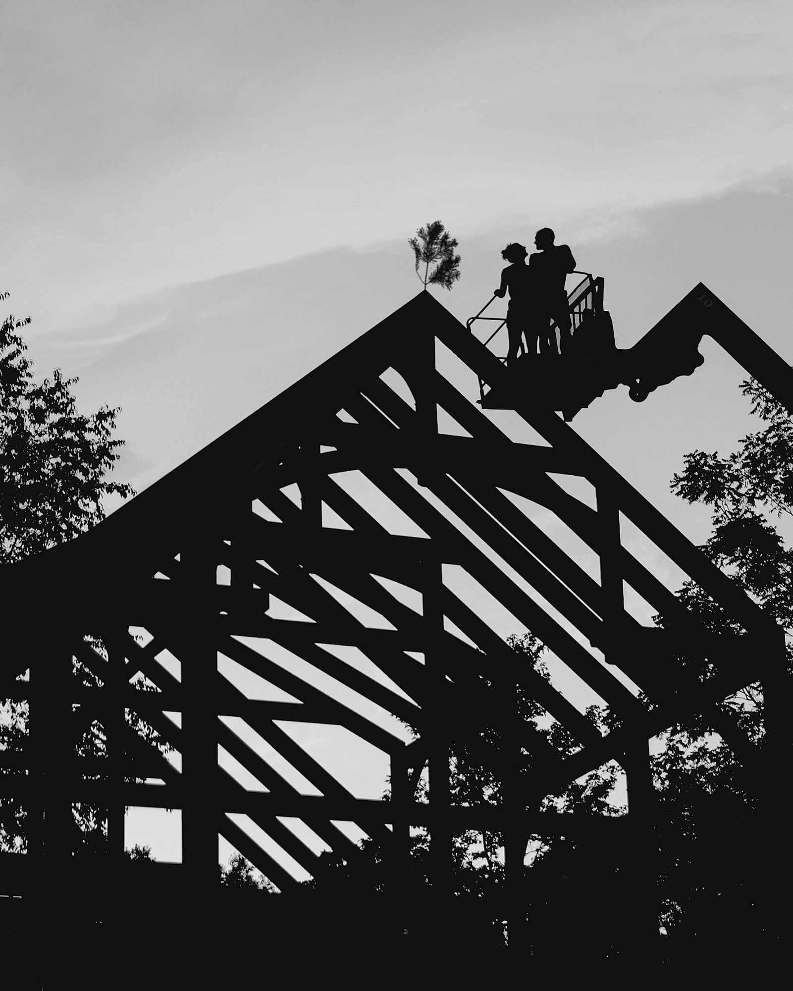 Two of the crew on a lift at the peak, setting the pine bough, the frame in silhouette against the sunset