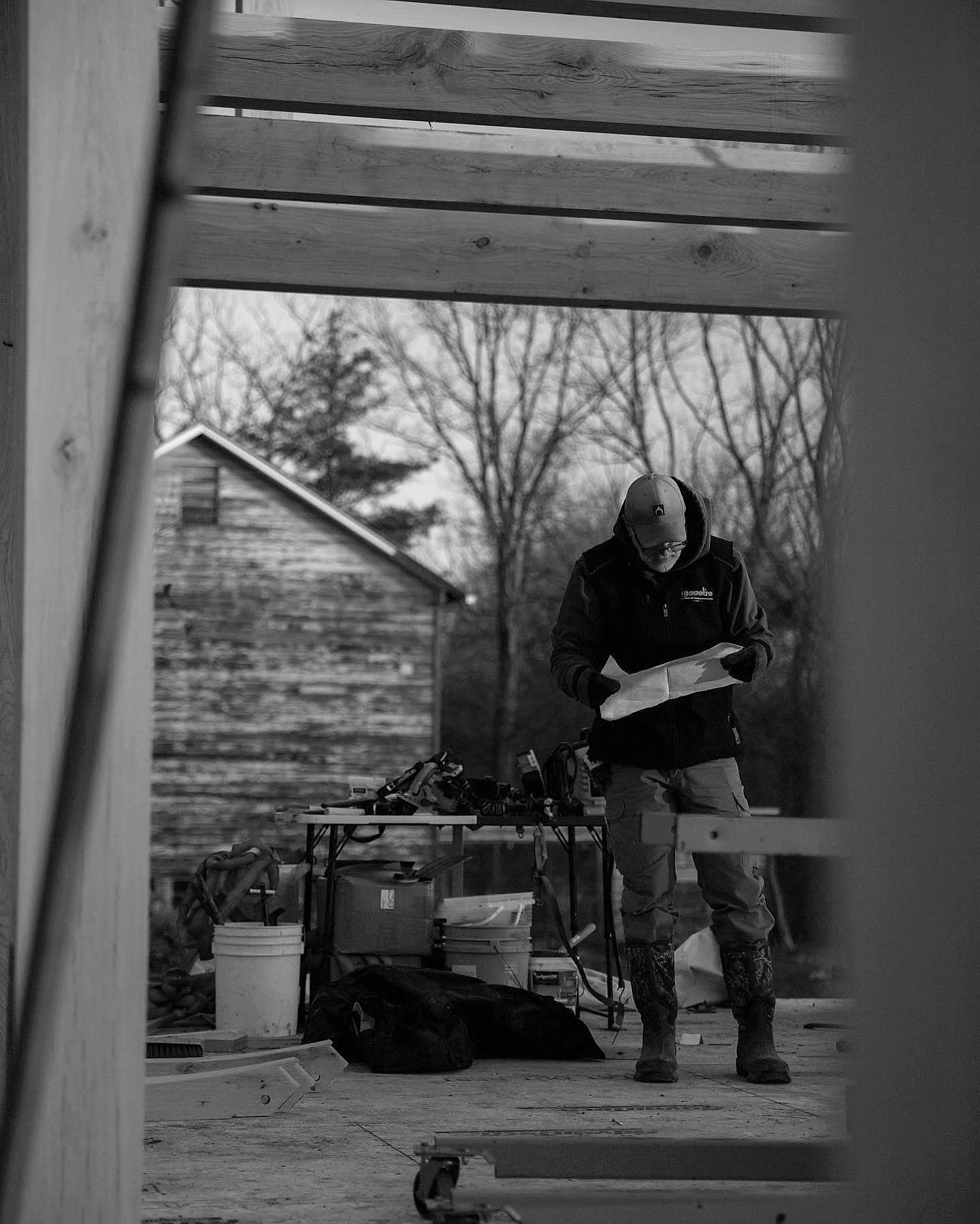 A framer reading the drawings on the deck at first light, seen through the frame