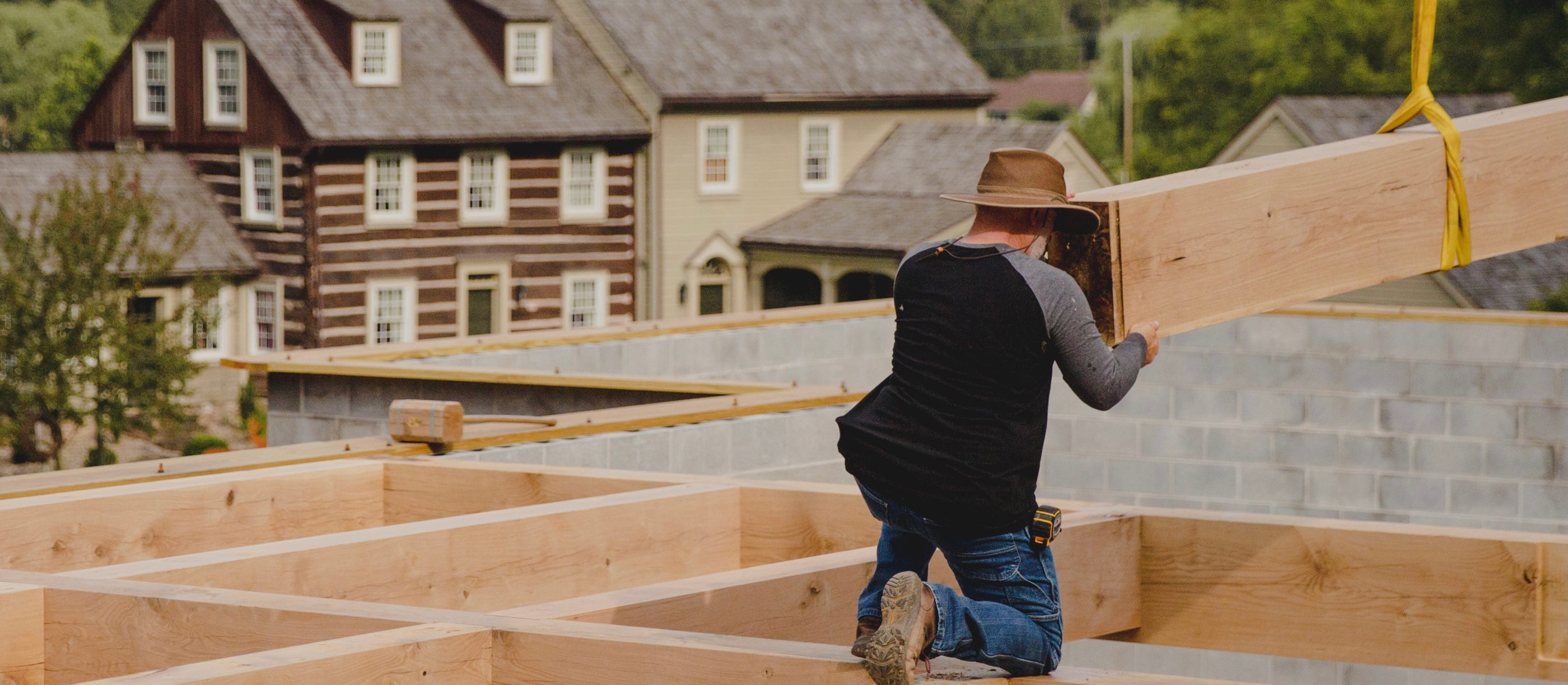 Crew member guiding a floor beam off the crane strap on site, a historic stone-and-log house on the hillside behind