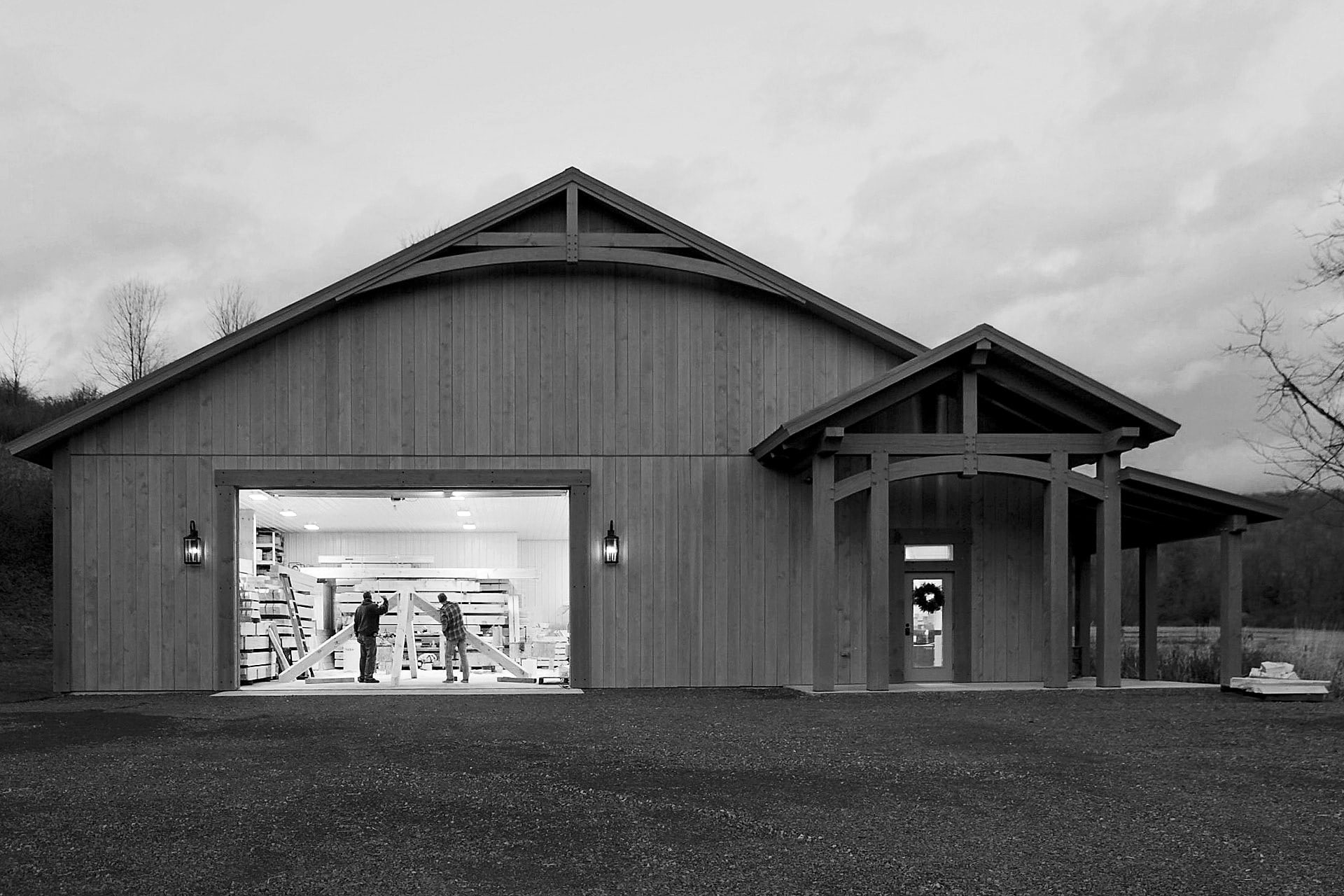 The shop at dusk, its big door open and two framers carrying a truss inside