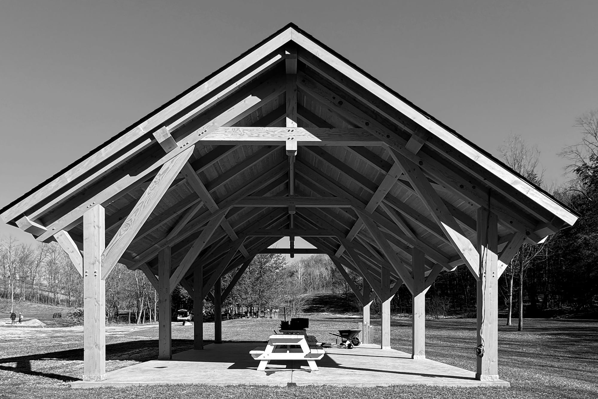 The finished pavilion under a blue sky, its timber frame beneath the roof, a picnic table on the slab, black and white
