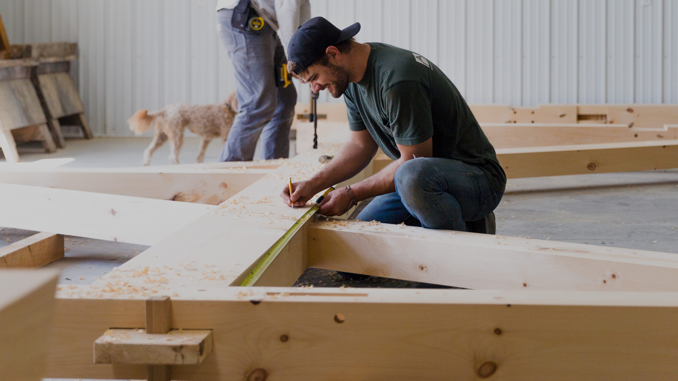 Framer kneeling on a test-fit truss in the shop, measuring and marking a joint amid loose shavings