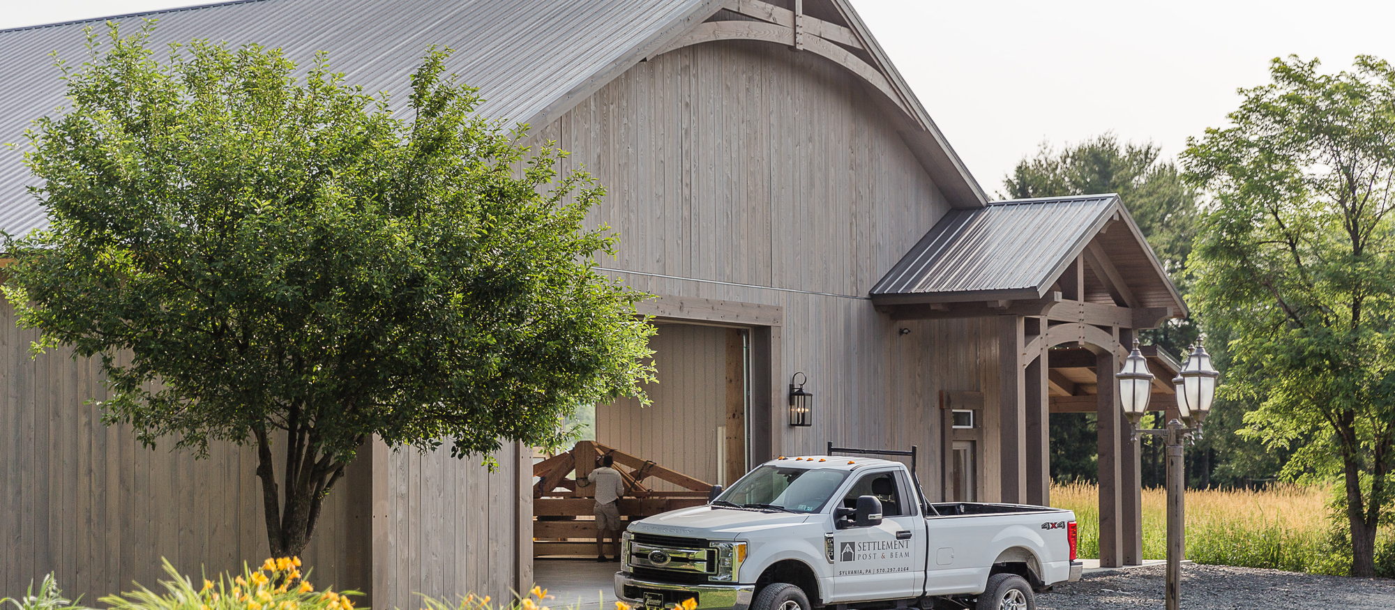 A Settlement Post & Beam truck parked outside a board-and-batten barn with a timber frame entry porch, someone at work just inside the open door