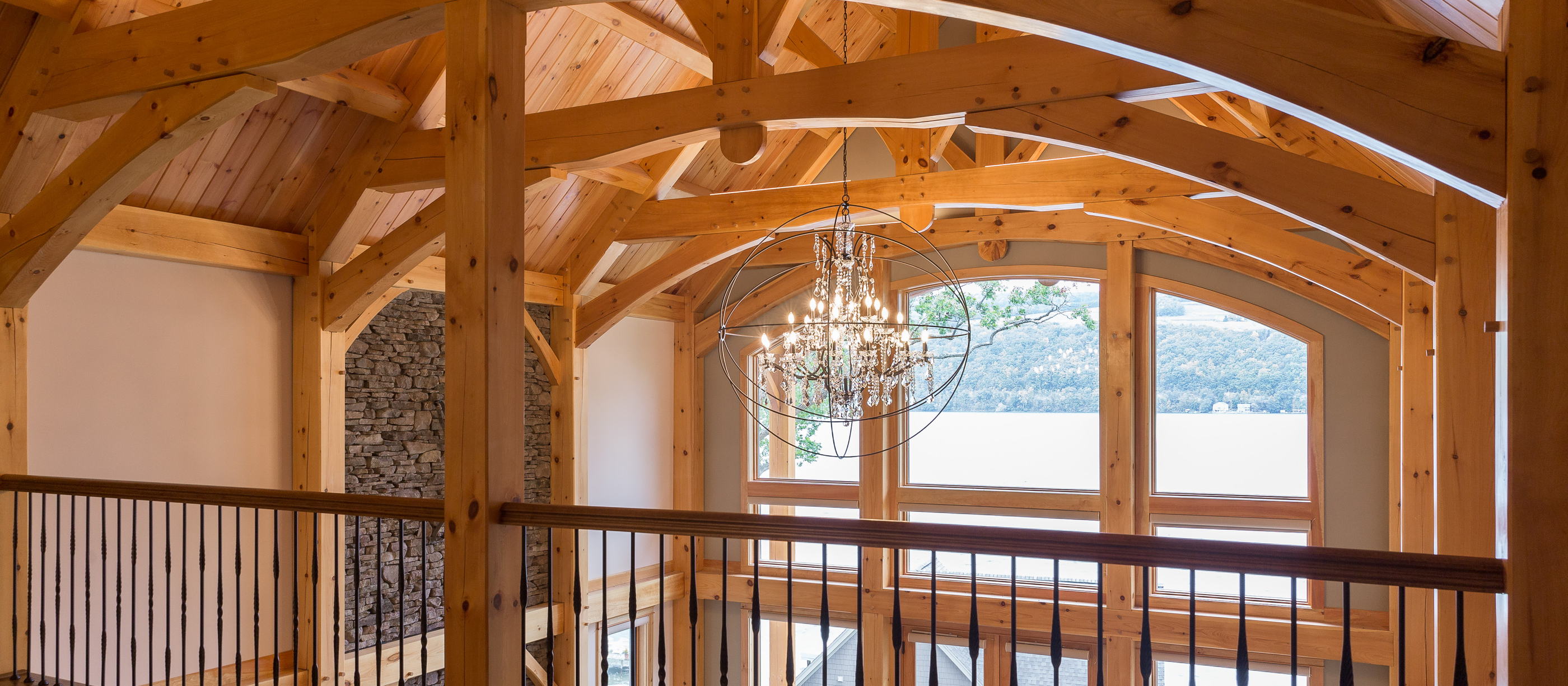 A great room under arched king post trusses, seen from the loft, the lake through the arched windows